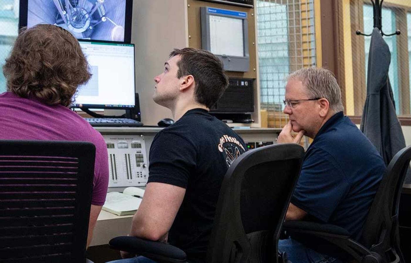 Three people sit in chairs and look at control systems of a nuclear reactor.