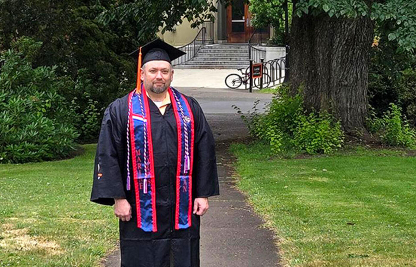 A person stands outside and looks toward the camera wearing academic regalia.