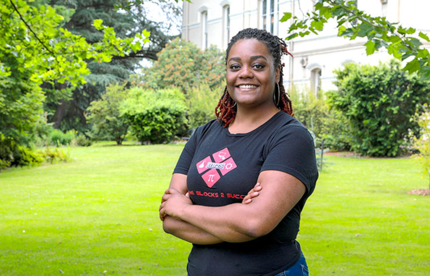 A person stands outside and smiles toward the camera with green grass and a historic building in the background.