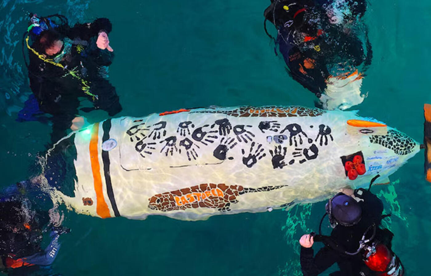 Four people in scuba diving gear float on the surface of water and touch a small submarine with the word Castorea painted on its side.