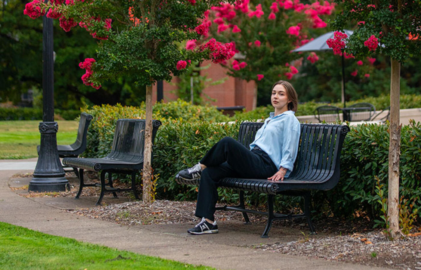 A person sits on a bench outside with summer blooms overhead and looks toward the camera.
