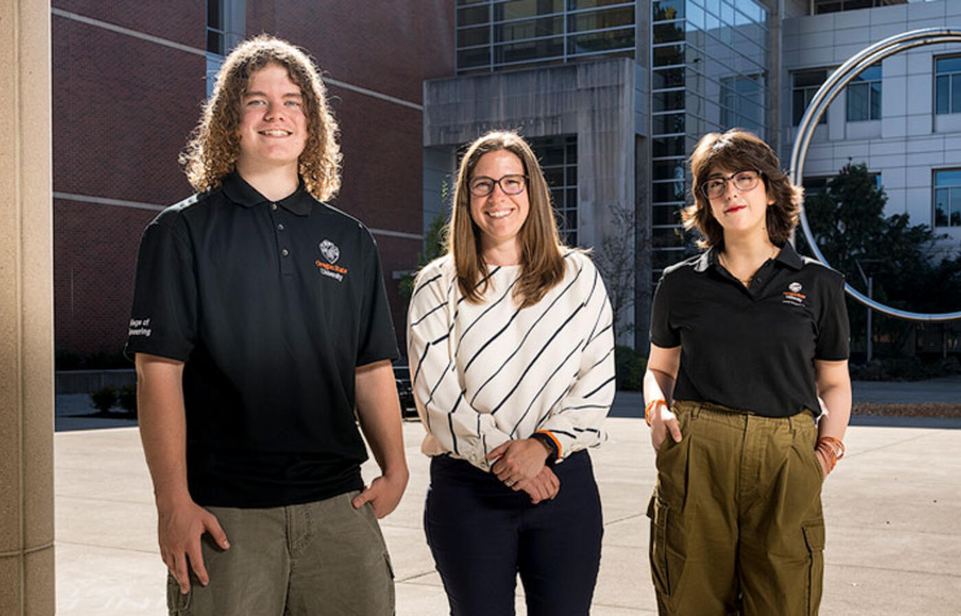 Three people stand outdoors on a campus with circular sculpture in the background and smile toward the camera. 