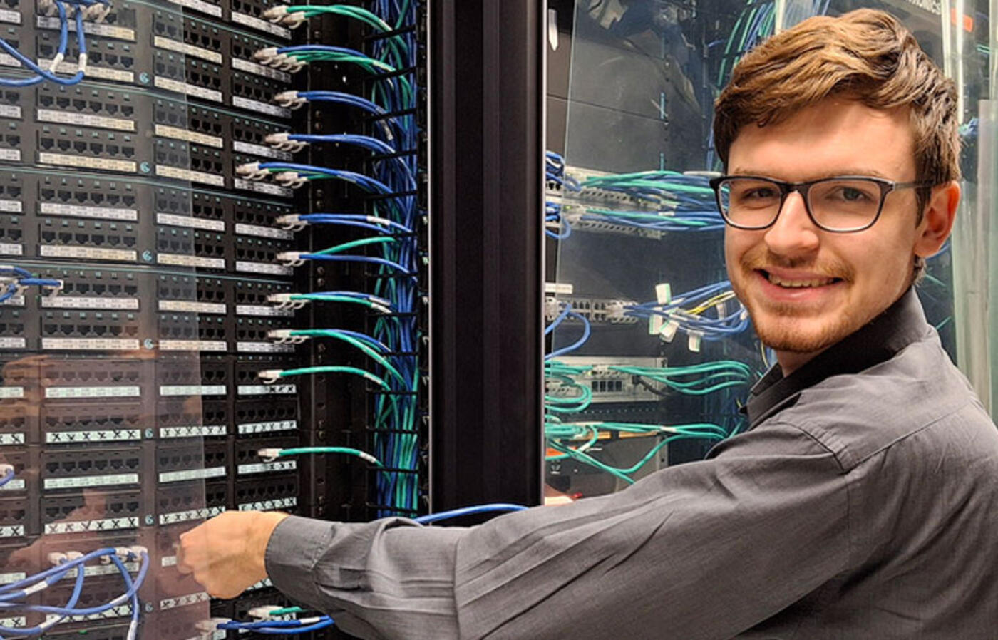 A person stands in a data center and adjusts hardware while looking toward the camera and smiling.