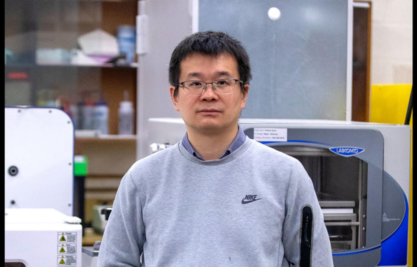 A person wears a light grey sweatshirt and stands in front of industrial equipment in a lab and looks toward the camera.