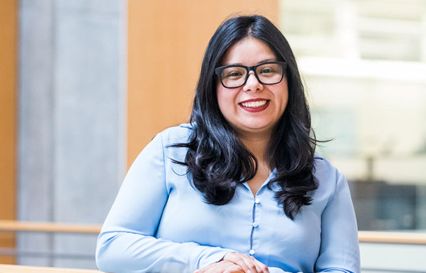 A person stands indoors near a wooden railing with a modern office background featuring glass panels and neutral-colored walls.