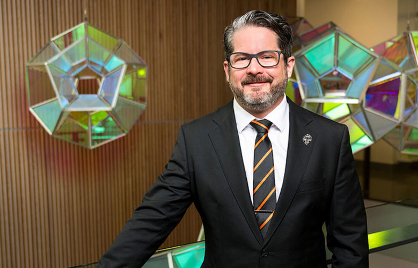 A person in a suit stands near a railing with a glass art installation behind.