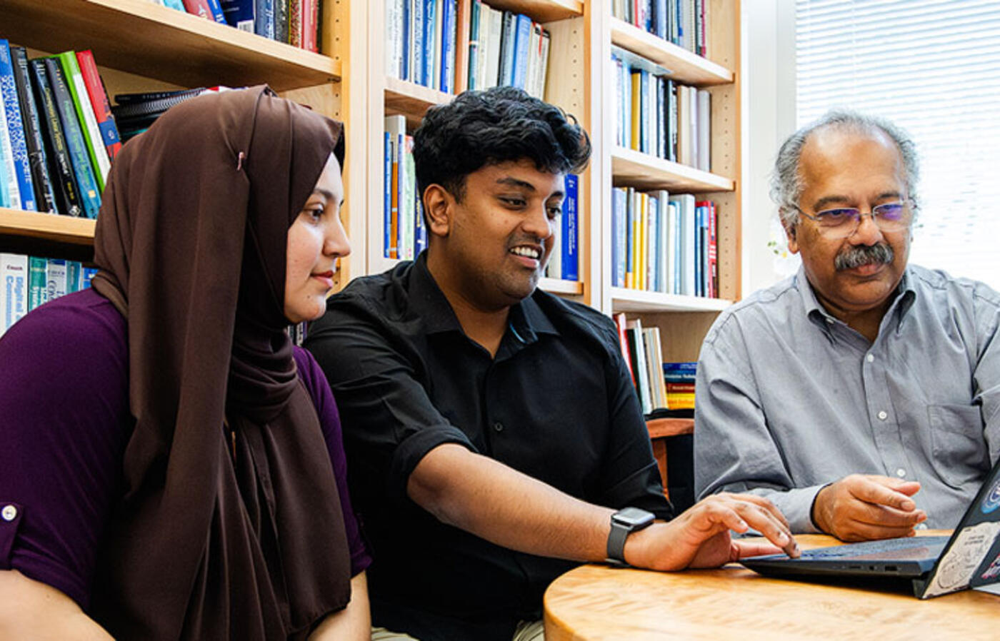 Three people sit at a table in an office and look at a laptop. 