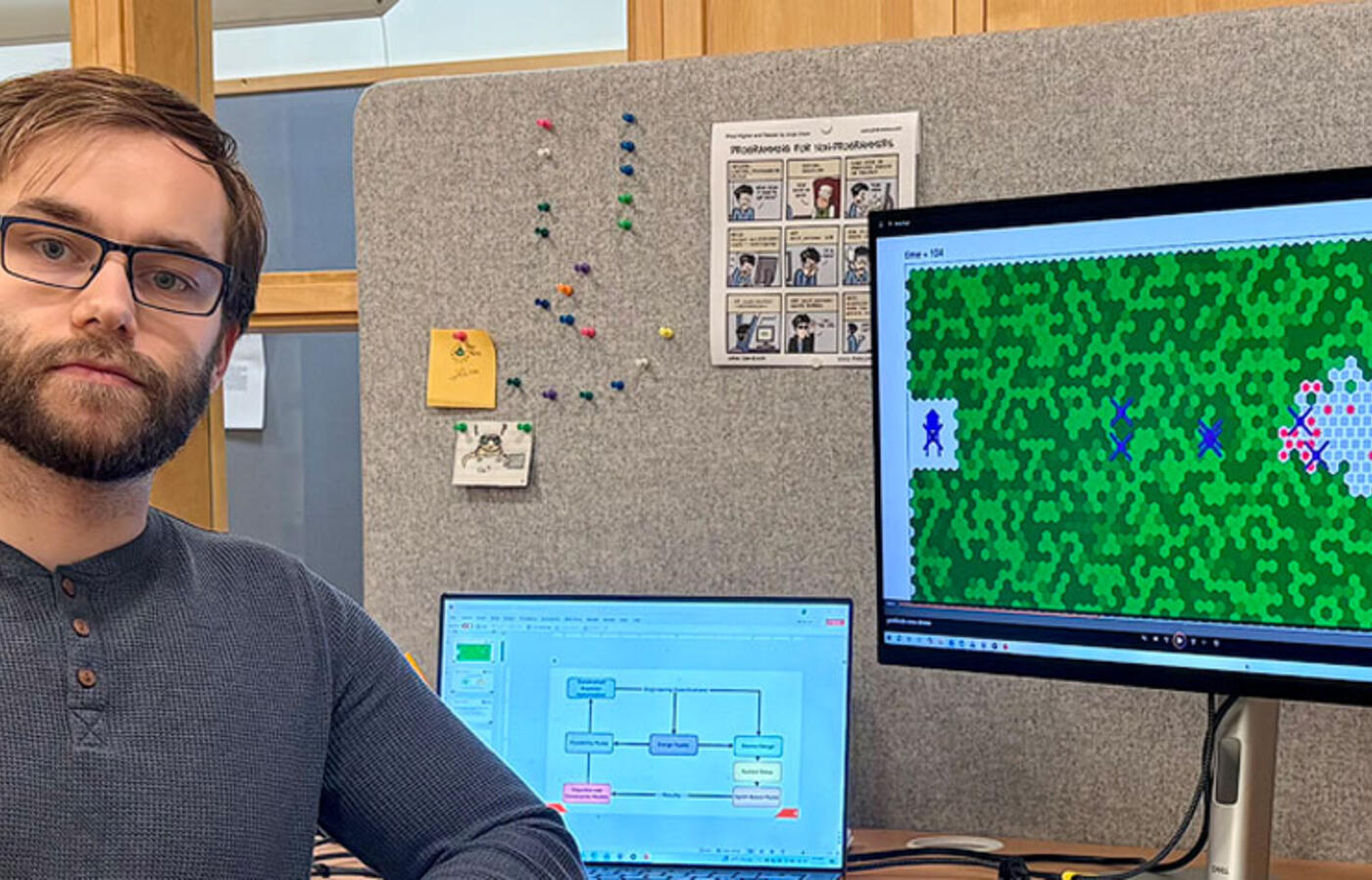 Person seated at a desk with two monitors; the larger monitor displays a green grid-based game with colored characters, while the smaller laptop screen shows a flowchart diagram.