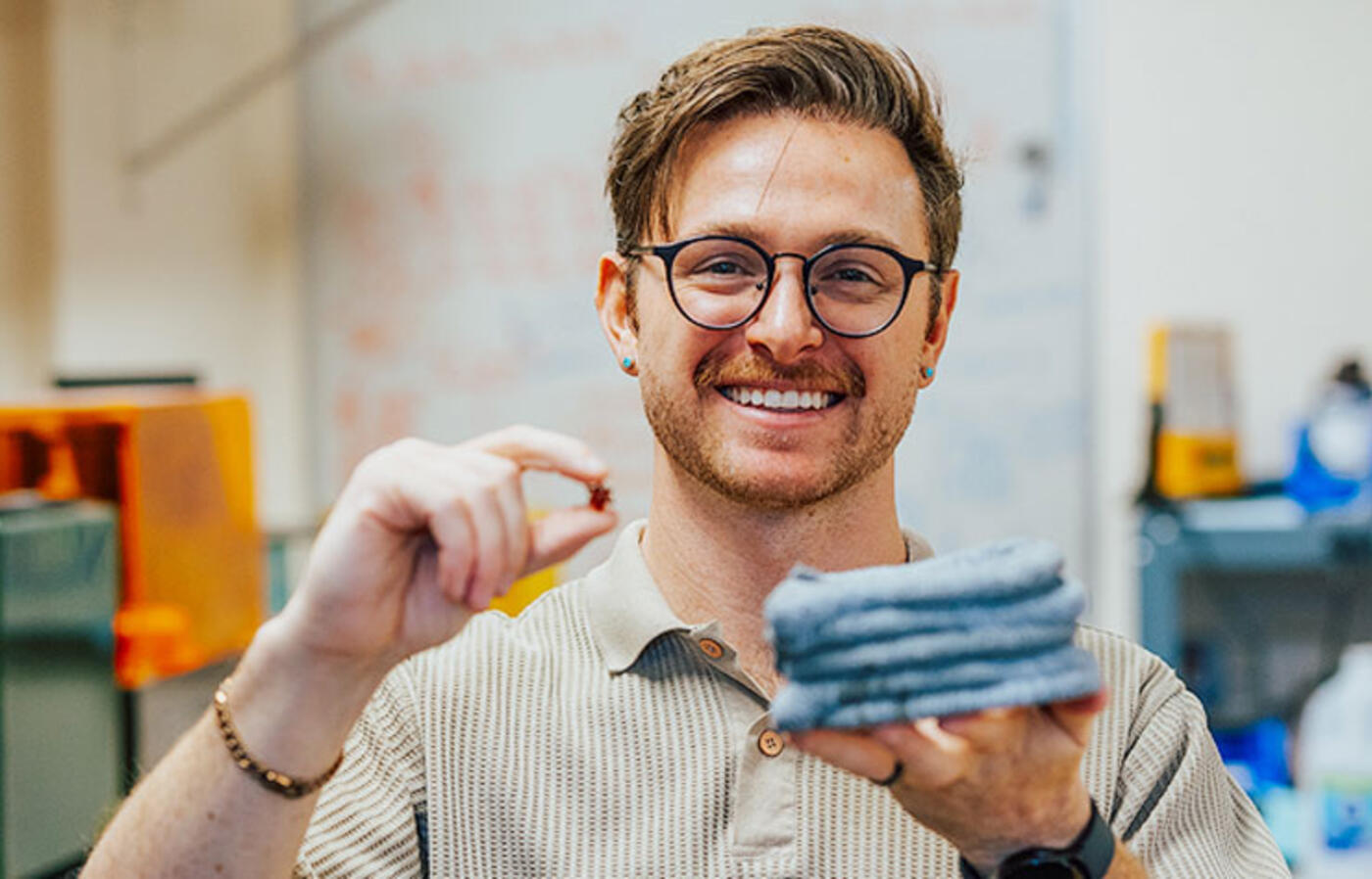 A person stands in a lab and holds a small item in their right hand and a stack of flat blue gray materials in their left hand and smiles toward the camera.