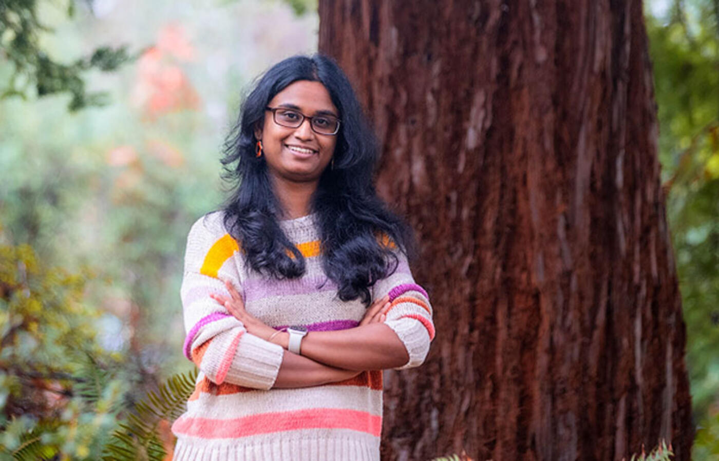 A person wears a striped sweater and stands outside in front of a large tree with green vegetation in the foreground.