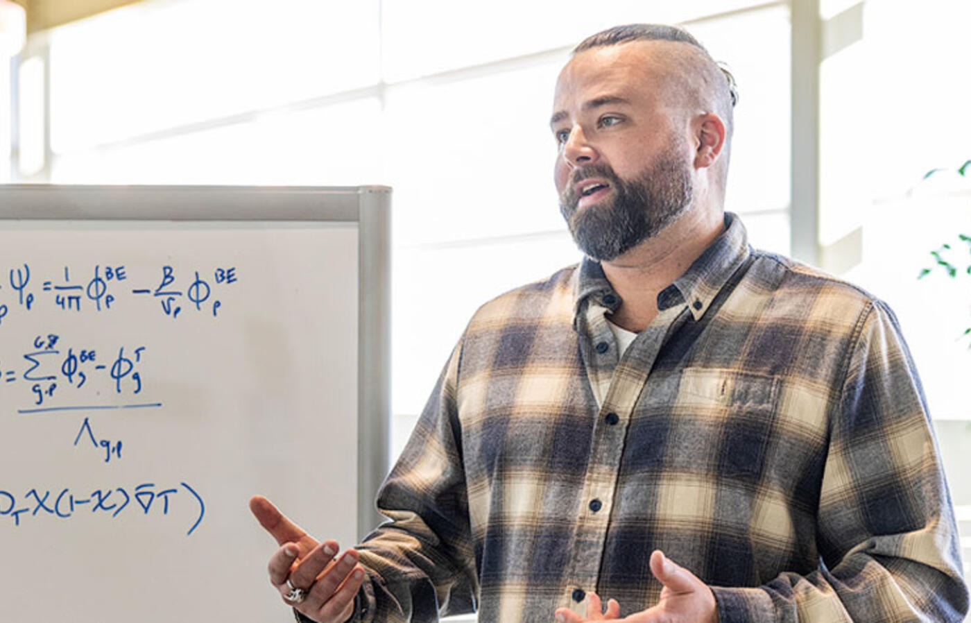 A person wears a plaid shirt and stands indoors near a whiteboard with complex mathematical equations.
