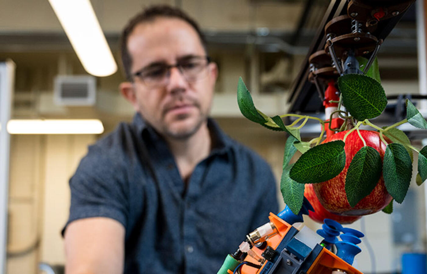 A person in a lab setting looks down at a robotic arm next to an apple. 