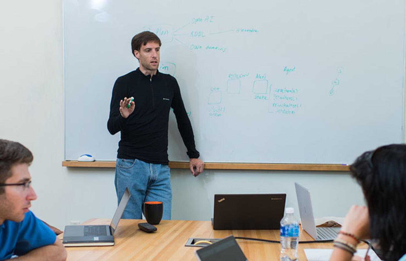 A person stands at the front of a conference room, pointing to a whiteboard covered with handwritten diagrams and notes, while several people sit around a table with laptops and notebooks, listening and discussing.