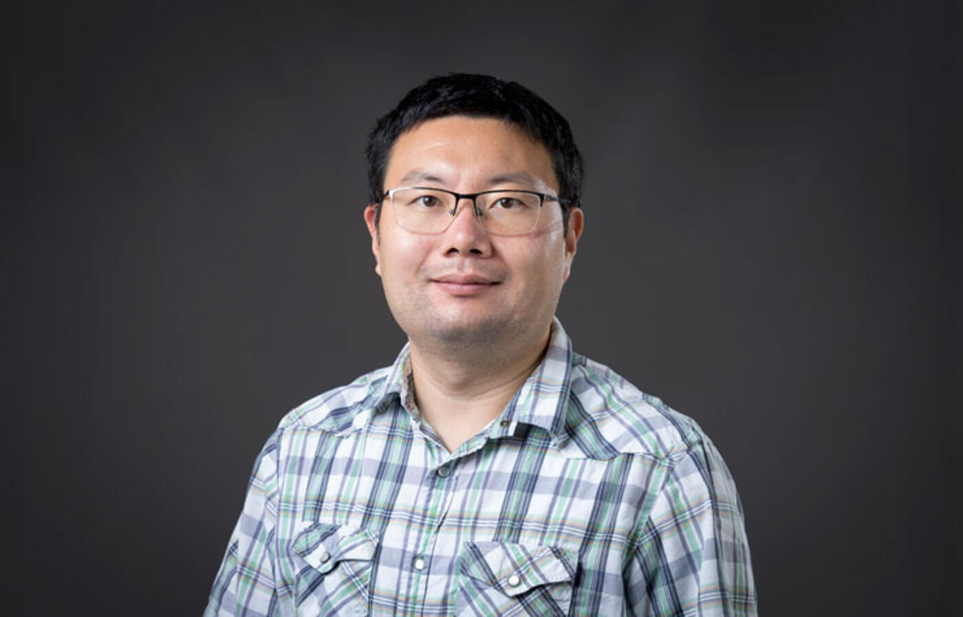 A person wears a light plaid button-up shirt and stands against a plain dark background, facing the camera.