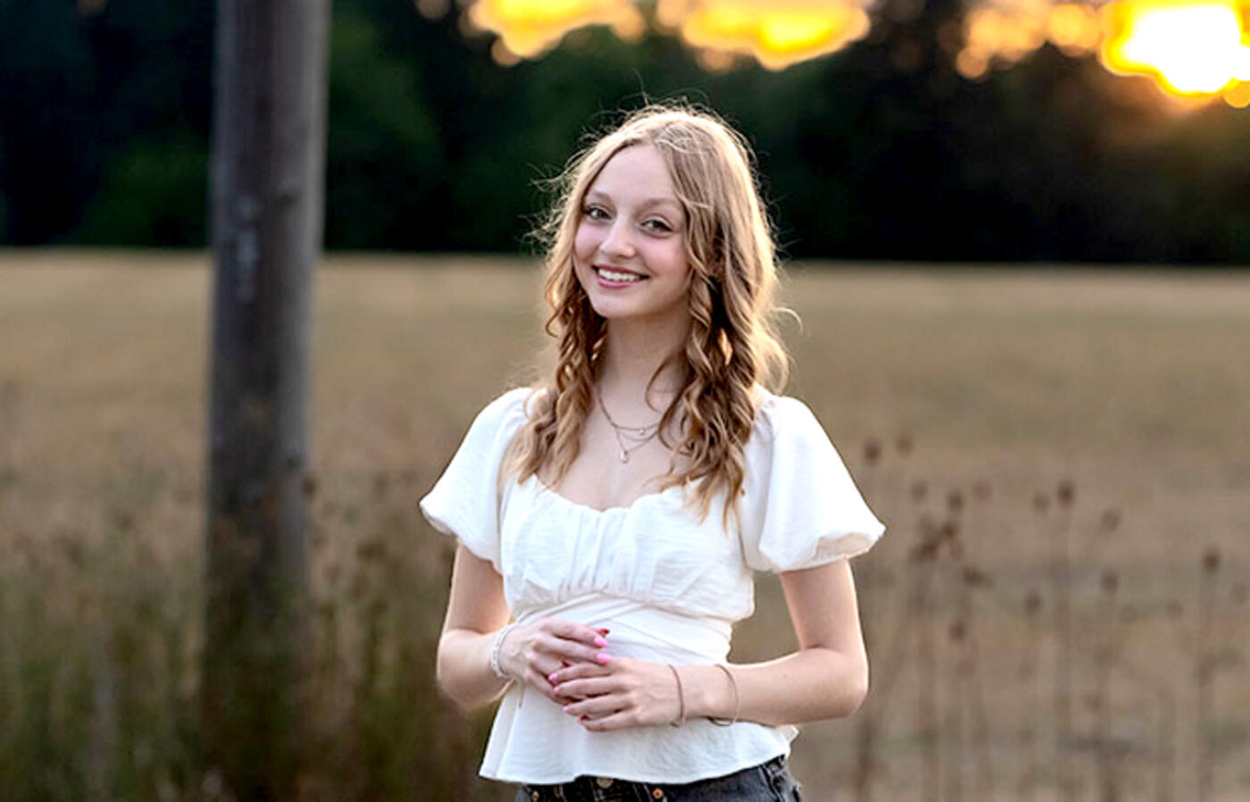 A person with shoulder-length hair stands in a field at sunset and wears a white short-sleeve shirt. 