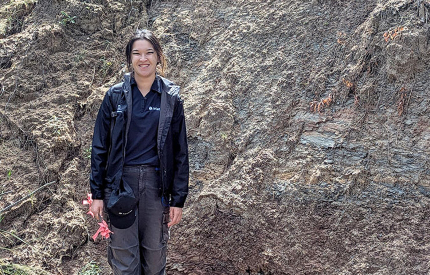 A person stands outdoors beside an exposed rock wall with sparse vegetation and small trees around them.