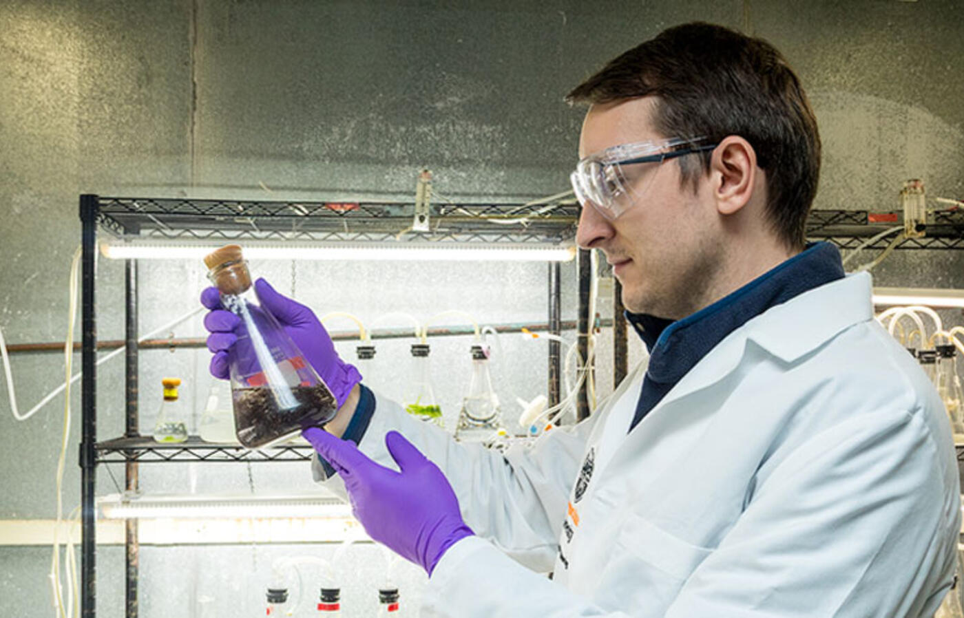 A person in a lab wears a lab coat and gloves while holding and examining a glass flask containing a dark liquid, standing in front of metal shelving.