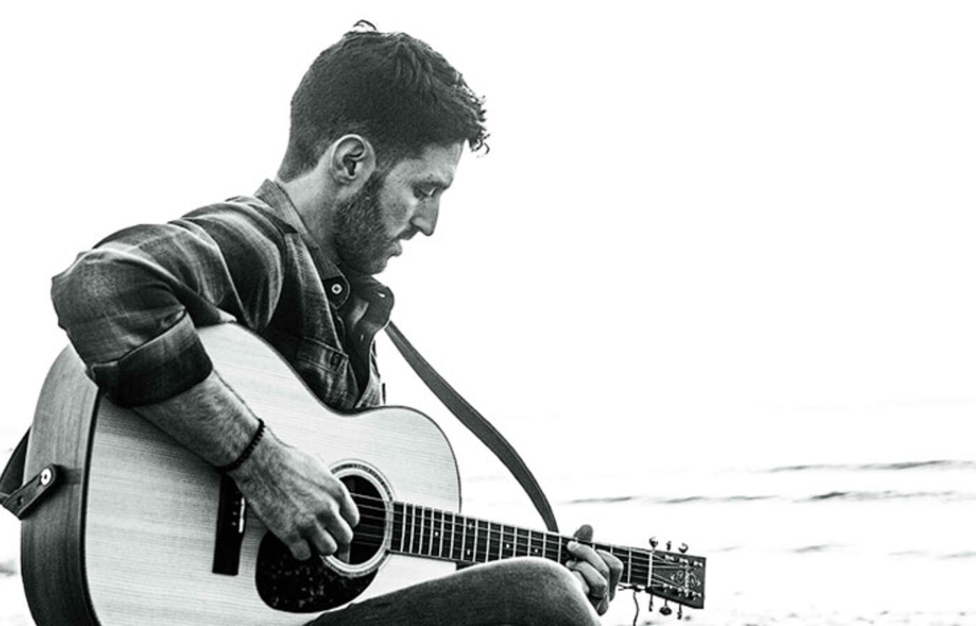 A person sits on a beach, playing an acoustic guitar with ocean waves and distant rocks in the background.