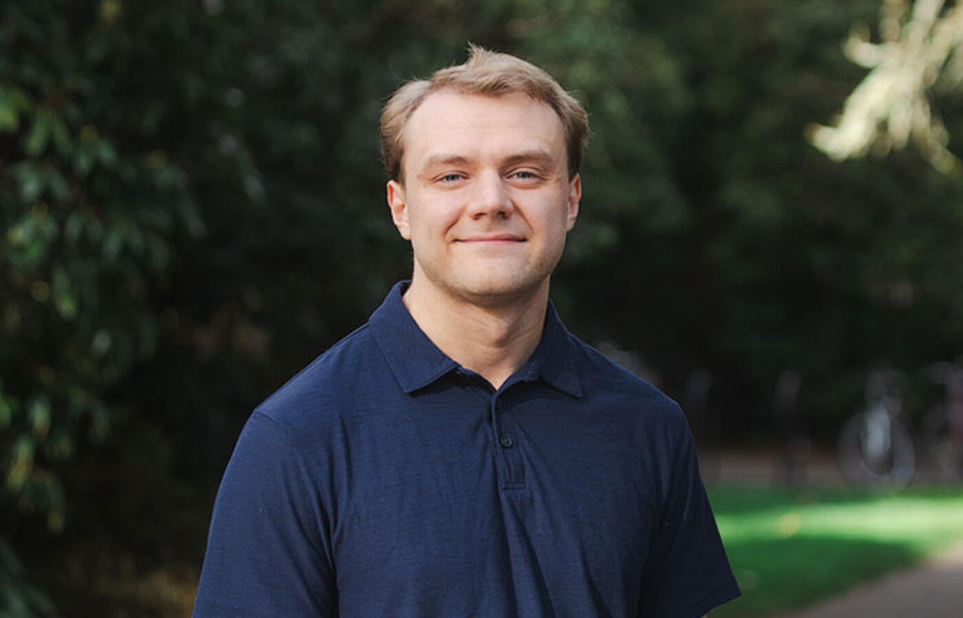 A person stands on a paved path with green grass and trees and looks toward the camera.