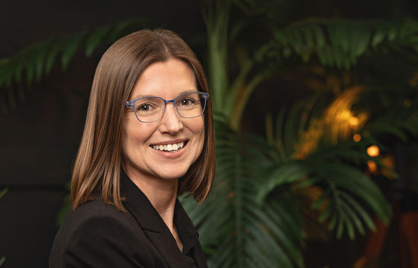 Camille Palmer wears business attire and sits in a dark area with tropical vegetation in the background and smiles toward the camera. 