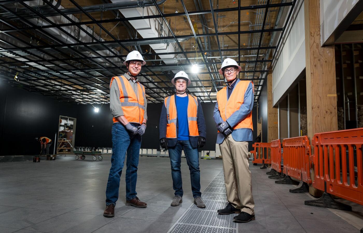 Anthony Koppers, Shayne Huddleston, and Todd Palmer dressed in hard hats and safety vests in a dark room under construction.