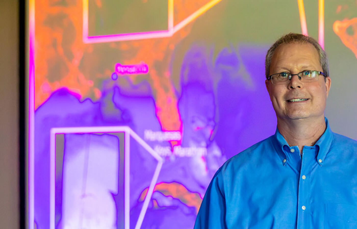 A person in business attire stands in a classroom with a digital projection of colorful bathymetry images behind them.