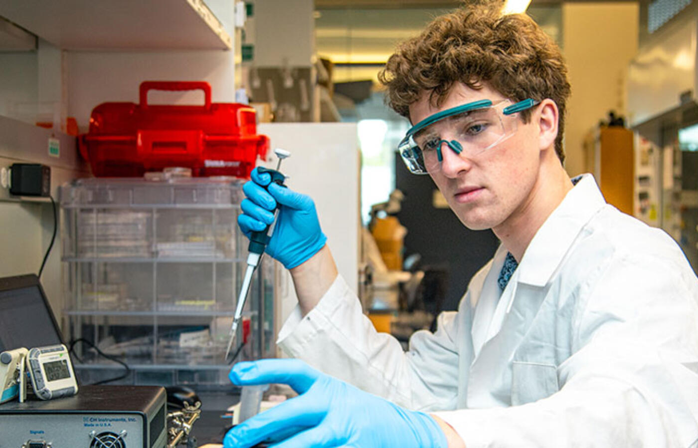 A man in a white lab coat sits at a lab bench, handling equipment and taking notes.