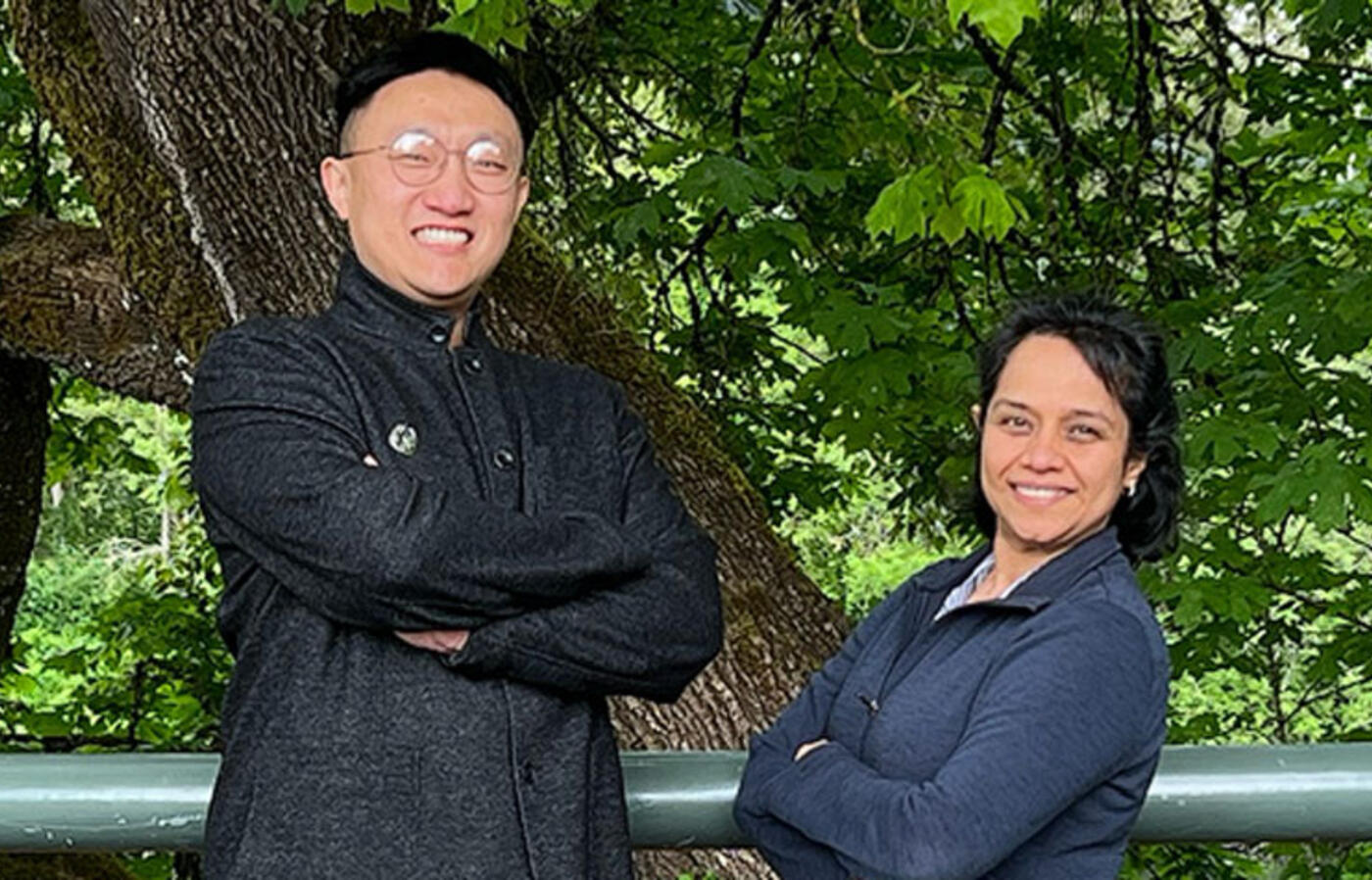 Two people wear black and stand outside in front of vegetation and smile toward the camera.