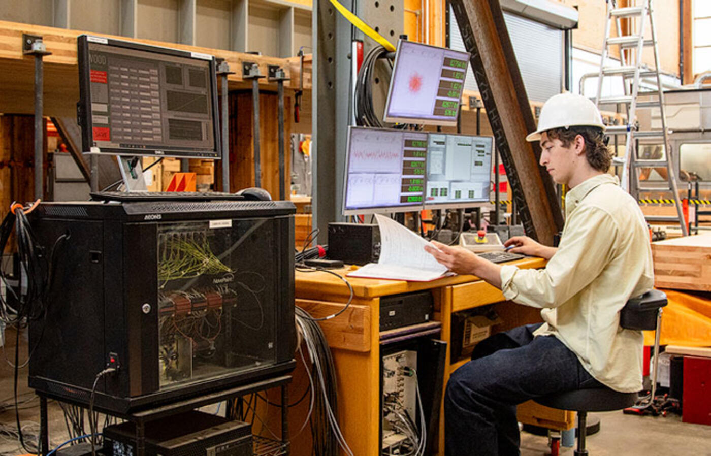 Dylan Goff wears a hard hat and sits in front of a computer and monitoring equipment in the Emmerson Lab, a large timber engineering facility.