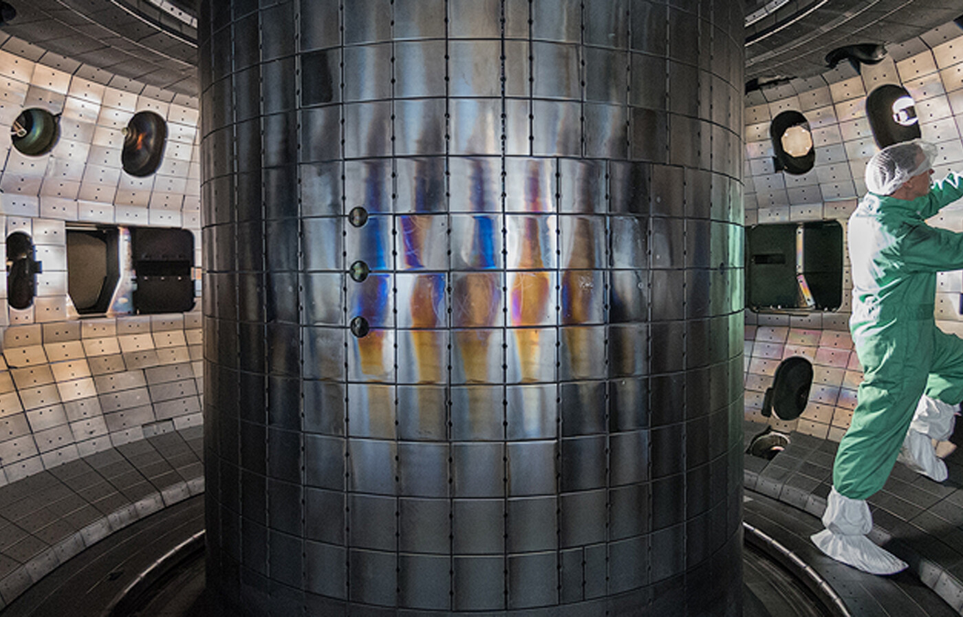 A person wears a clean-room suit with hair covering, gloves, and boot covers and stands inside a large cylindrical chamber lined with metallic panels and equipment.