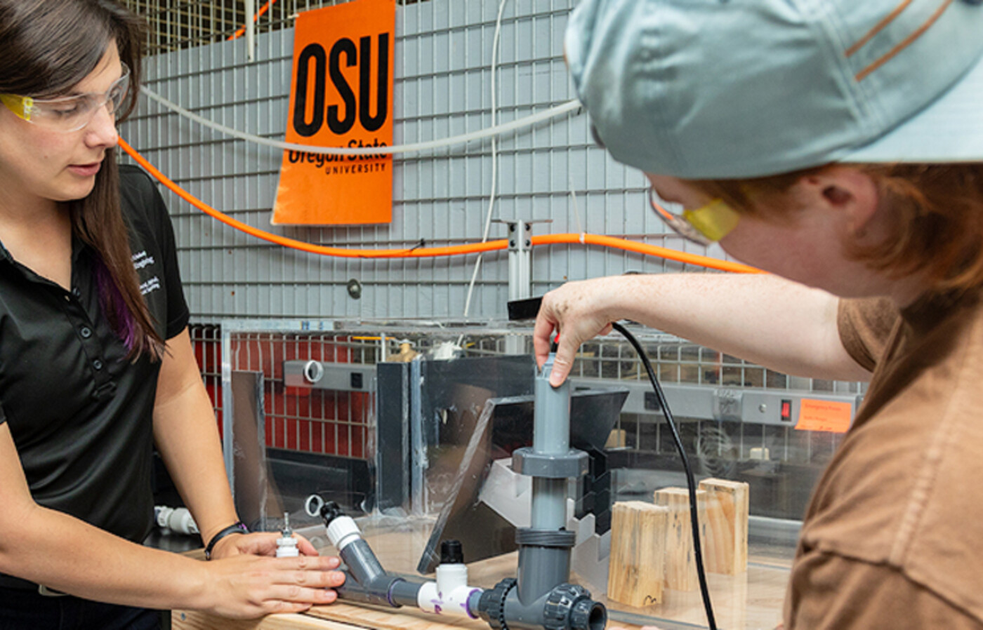 Kathryn Newhart and Perry Wilson wear eye protection and stand next to a workbench in a lab and adjust a device with two intersecting pipes.