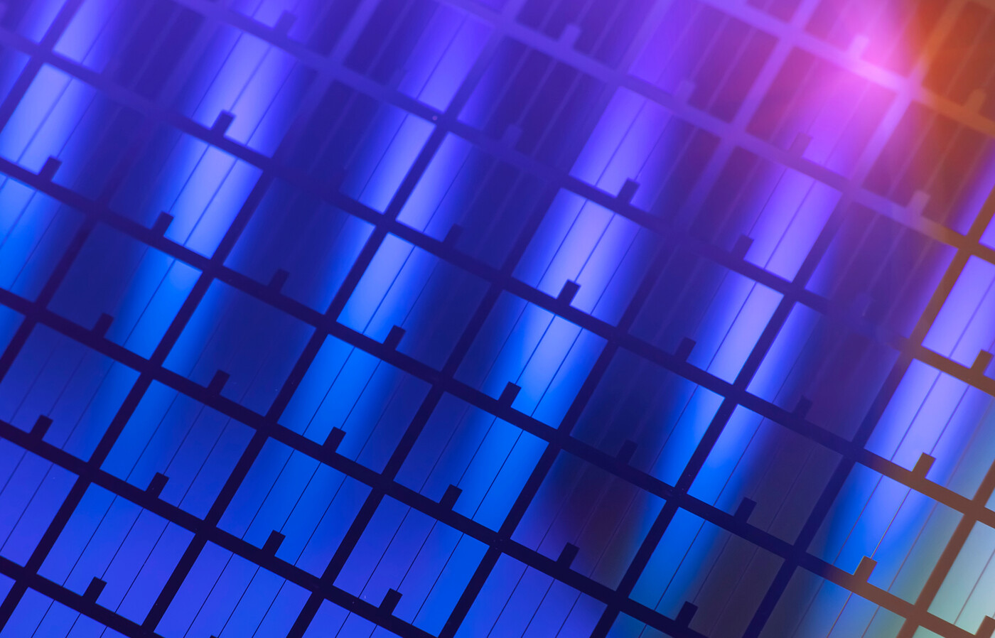 A close-up view of a semiconductor wafer with a repeating grid pattern of microelectronic structures in iridescent colors.