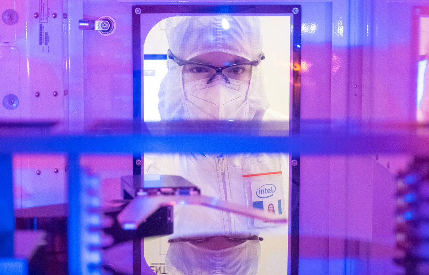 A person wears clean-room protective clothing, safety glasses, and a face mask is viewed through the interior of semiconductor manufacturing equipment in purple and blue light. 