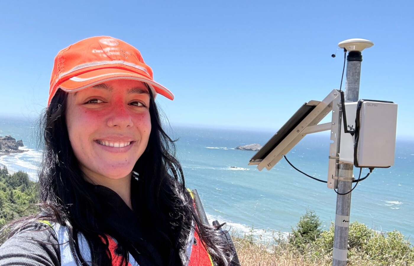 Heather Maran stands beside a coastal monitoring instrument overlooking the Oregon Coast with vegetation and ocean behind her.