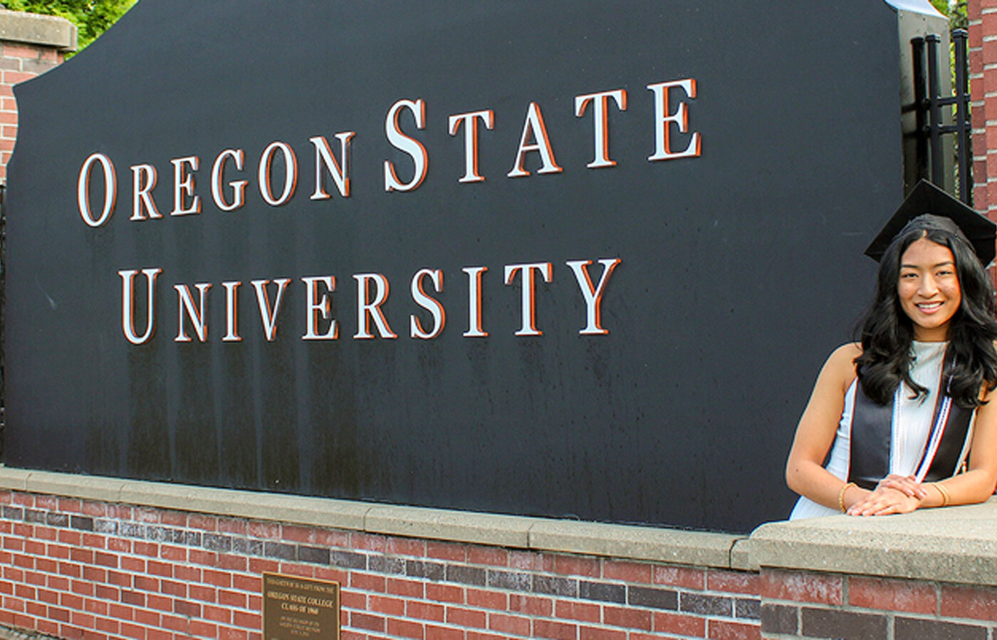 Megan Thomas wears a graduation cap and stole and stands outdoors leaning against a large black Oregon State University sign.