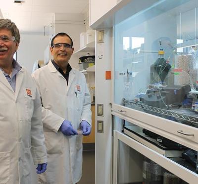 Two faculty member wearing lab coats in front of a protected glass with scientific materials inside it.