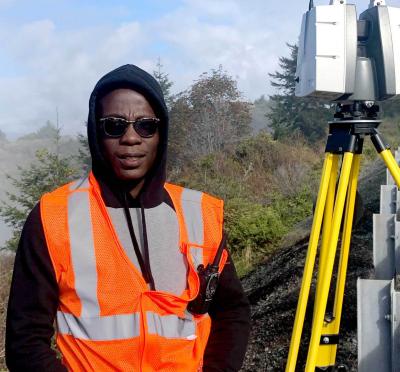 A student standing on the side of a coastal highway next to surveying equipment.