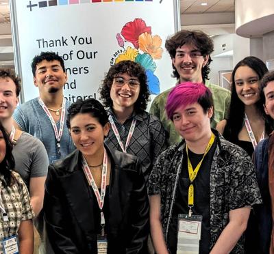 Nine students standing in front of a banner at a conference facility.