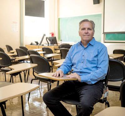 A guest instructor sitting at a desk in a classroom with chalkboard in the background.