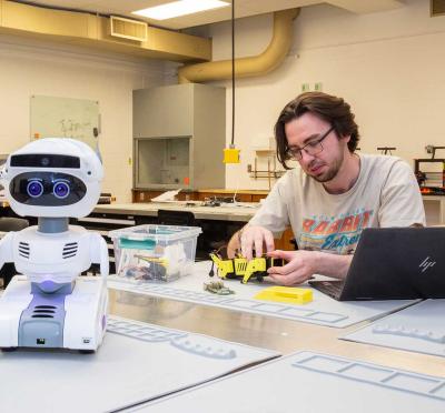 A student sitting at a workstation working on a small robot with a larger robot in the foreground. 