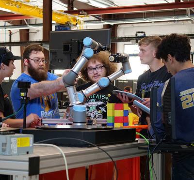 Eight students standing around a workstation at a lab with one student adjusting a robotic arm.