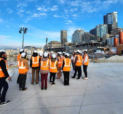 A group of students in personal protective equipment standing at a construction site near the water with the Seattle skyline in the background