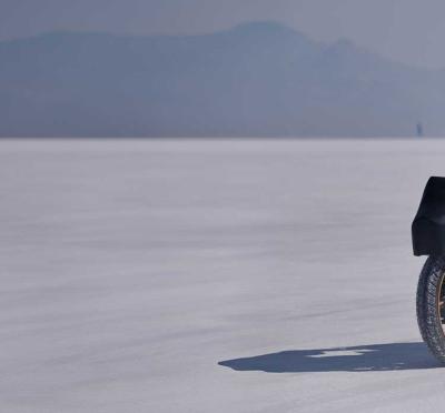 A motorcyclist riding a 50cc motorcycle across desert sand with mountains in the distance.