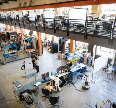 Two researchers standing in the center of a large open-room, multi-story research facility with robotics equipment and desks surrounding them. 