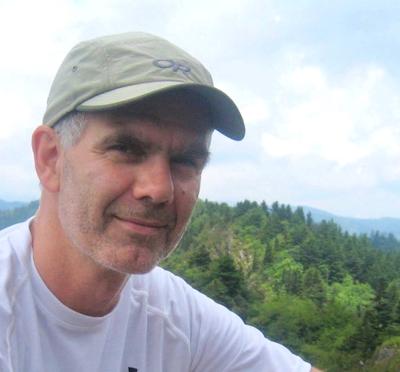 A person in a baseball cap sitting outside at a high vantage point with mountains in the background