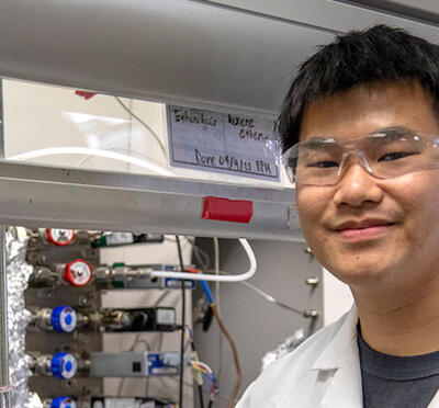 A person wearing a lab coat and eye protection stands in front of equipment in a lab facing the camera.