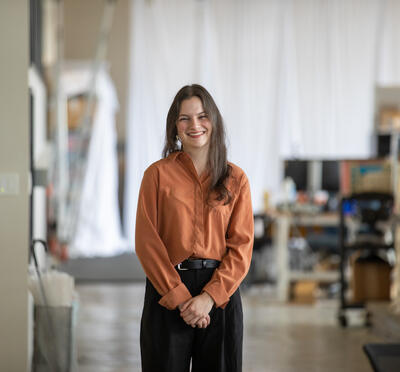Madelyn Sadler, a person wearing black pants and a rust colored top with long dark hair, clasps their hands in front of them while standing in a blurred-out workshop.