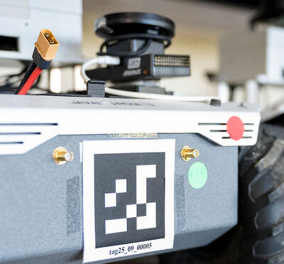 Two four-wheel silver and grey robot vehicles with black all-terrain tires sit on a shelf in a lab. They each have QR codes on the front, with wires. 