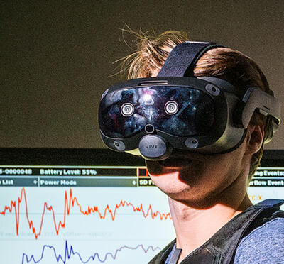 A person wears an augmented reality headset inside a lab and a digital screen displays data in the background.