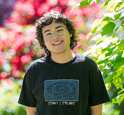 A person wearing a t-shirt smiles at the camera and stands in front of trees and spring blooms.
